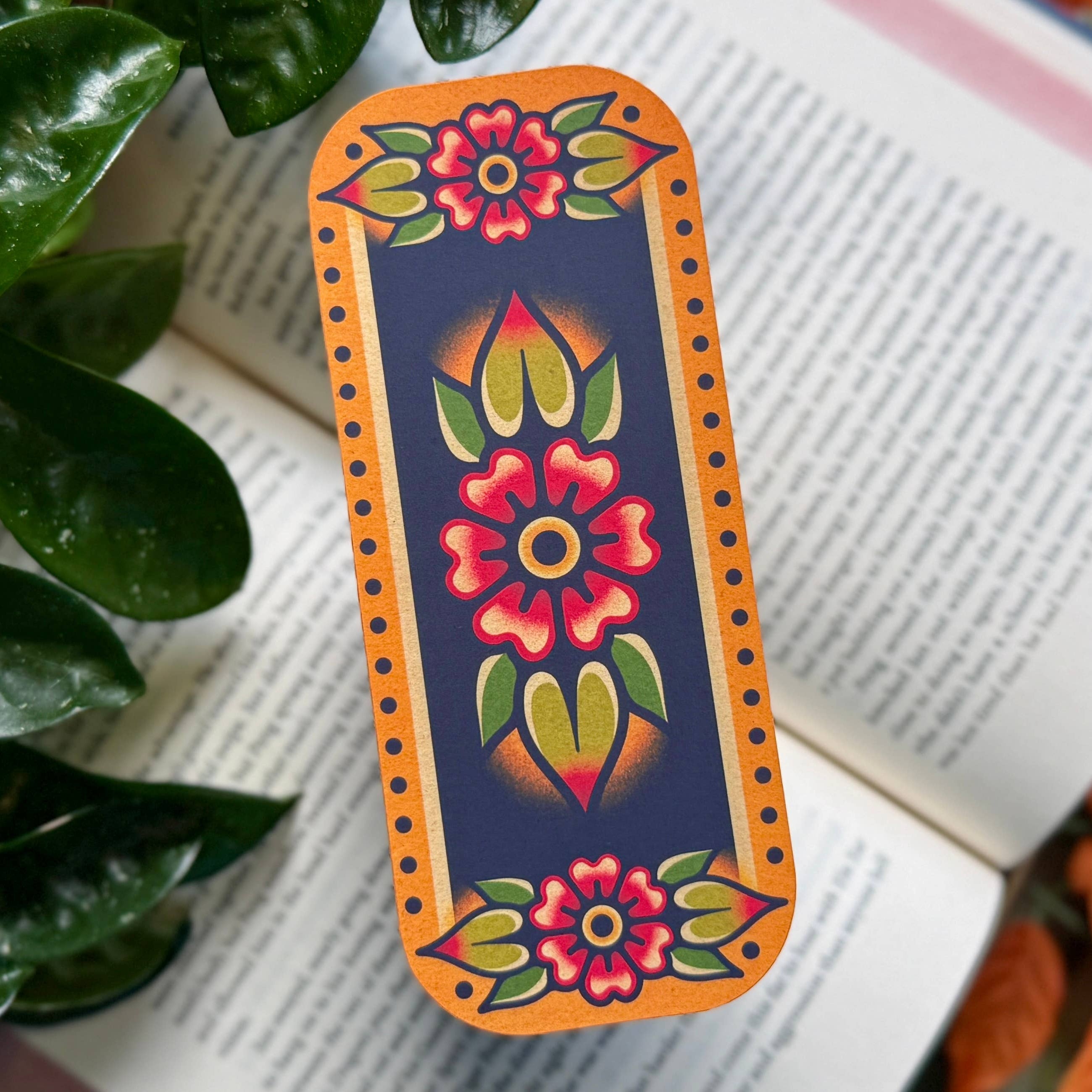 Tattoo-style flower bookmark with red and green floral designs on a navy background, photographed on an open book with greenery