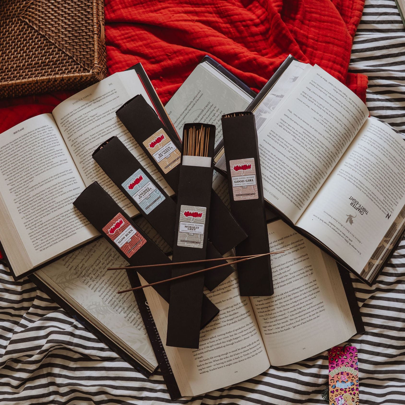 Flatlay of black incense sticks fanned across open romance books with striped bedding and red fabric underneath.