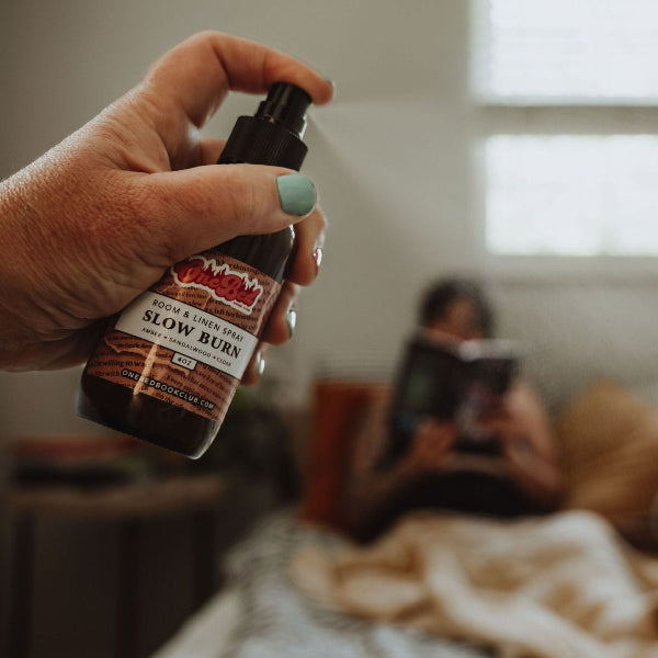 Person spraying room mist while sitting on bed surrounded by books and cozy pillows.