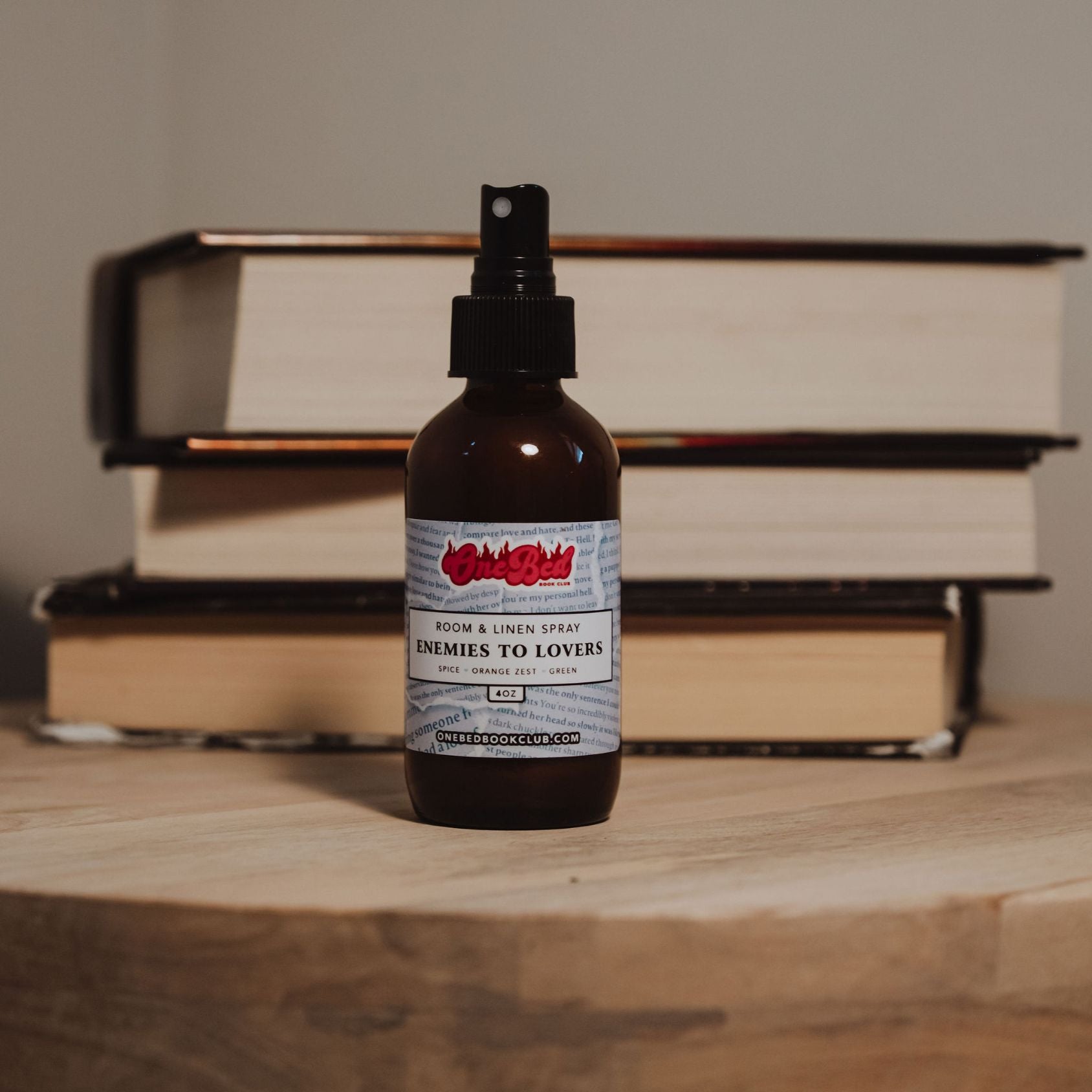 Close-up of One Bed Room Spray bottle titled “enemies to lovers” nestled among books.