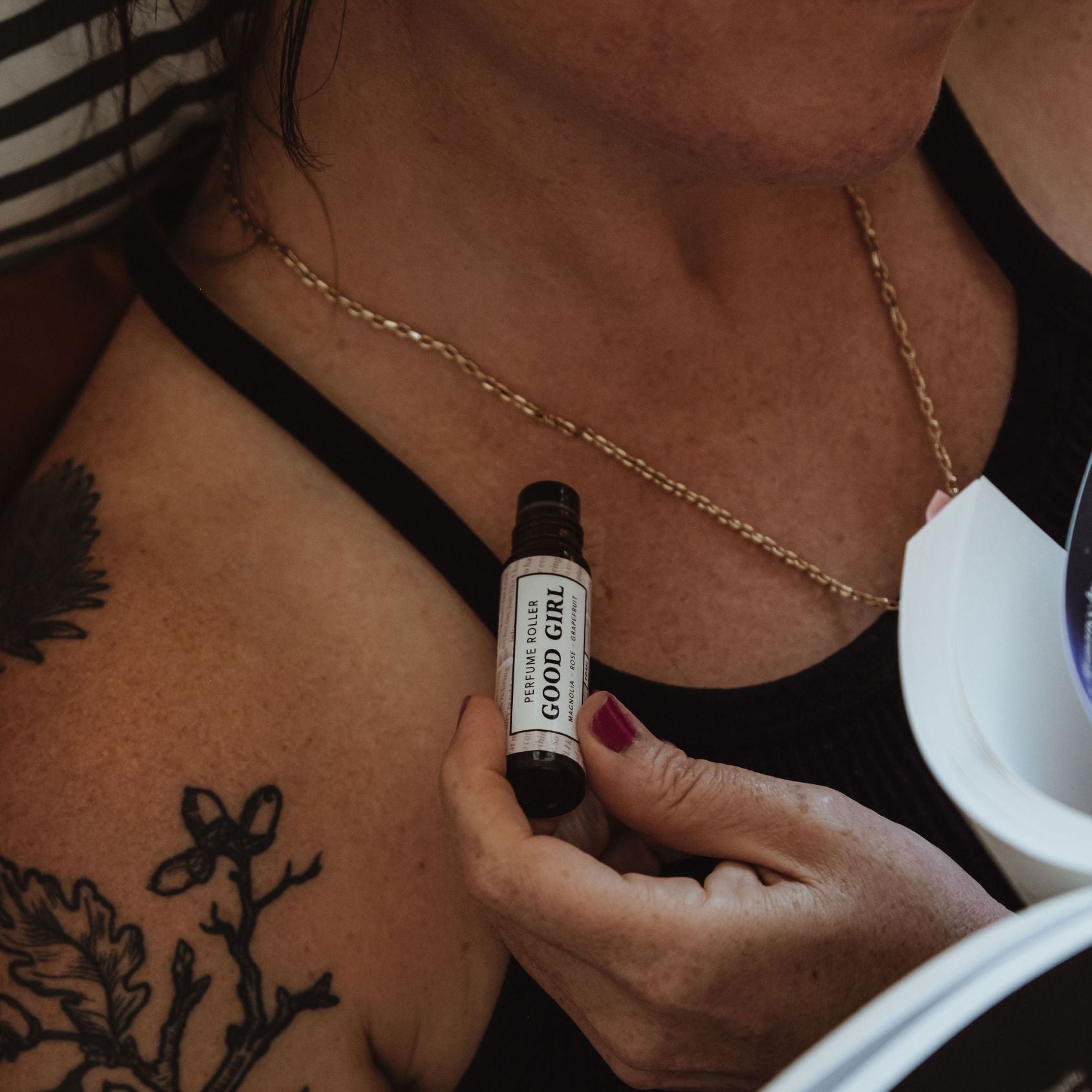 tattooed woman applying perfume roller to neck with books and striped bedding visible in background.