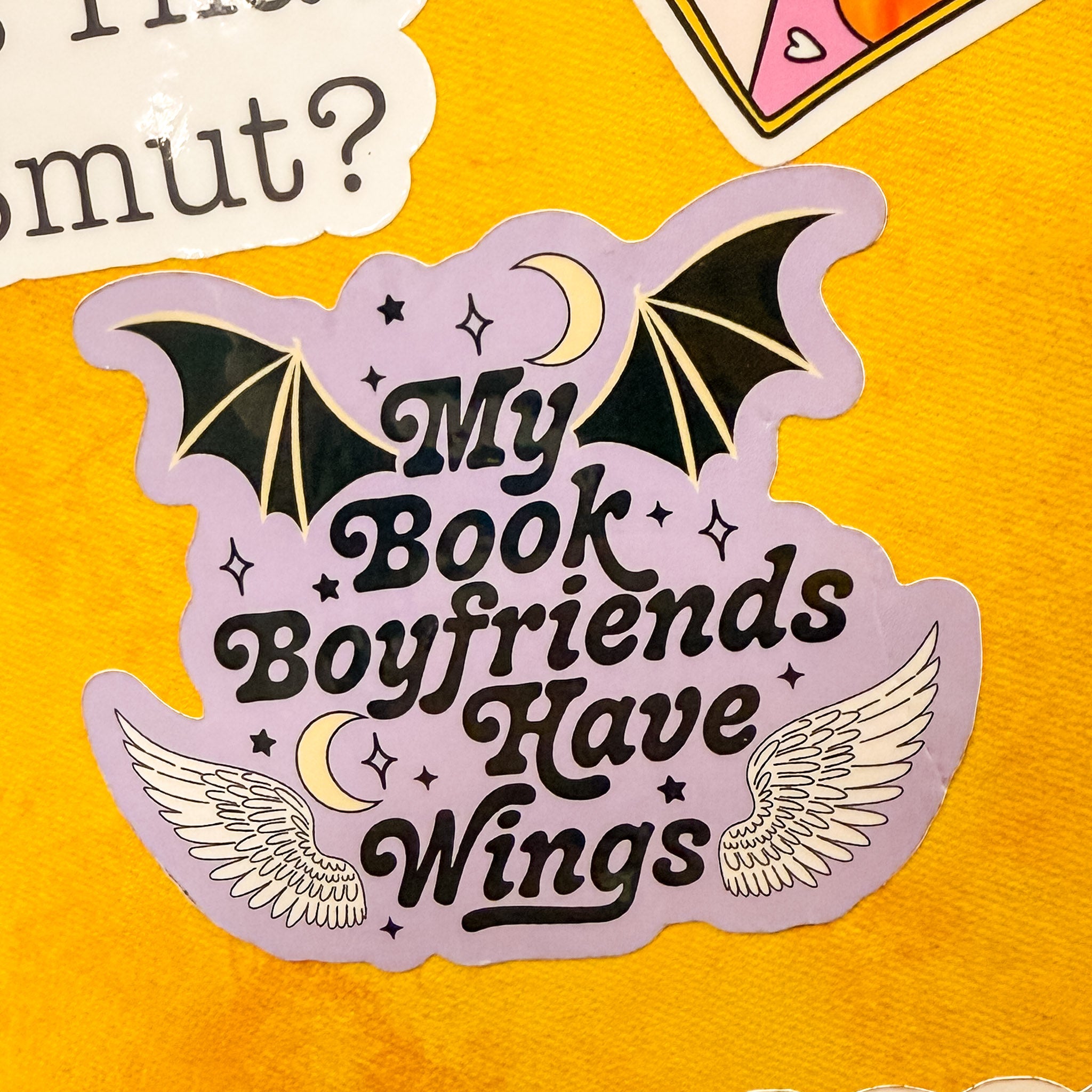 purple vinyl sticker that says "my book boyfriends have wings" with bat wings, stars and a crescent moon, shown on a bright yellow kindle case with other stickers.