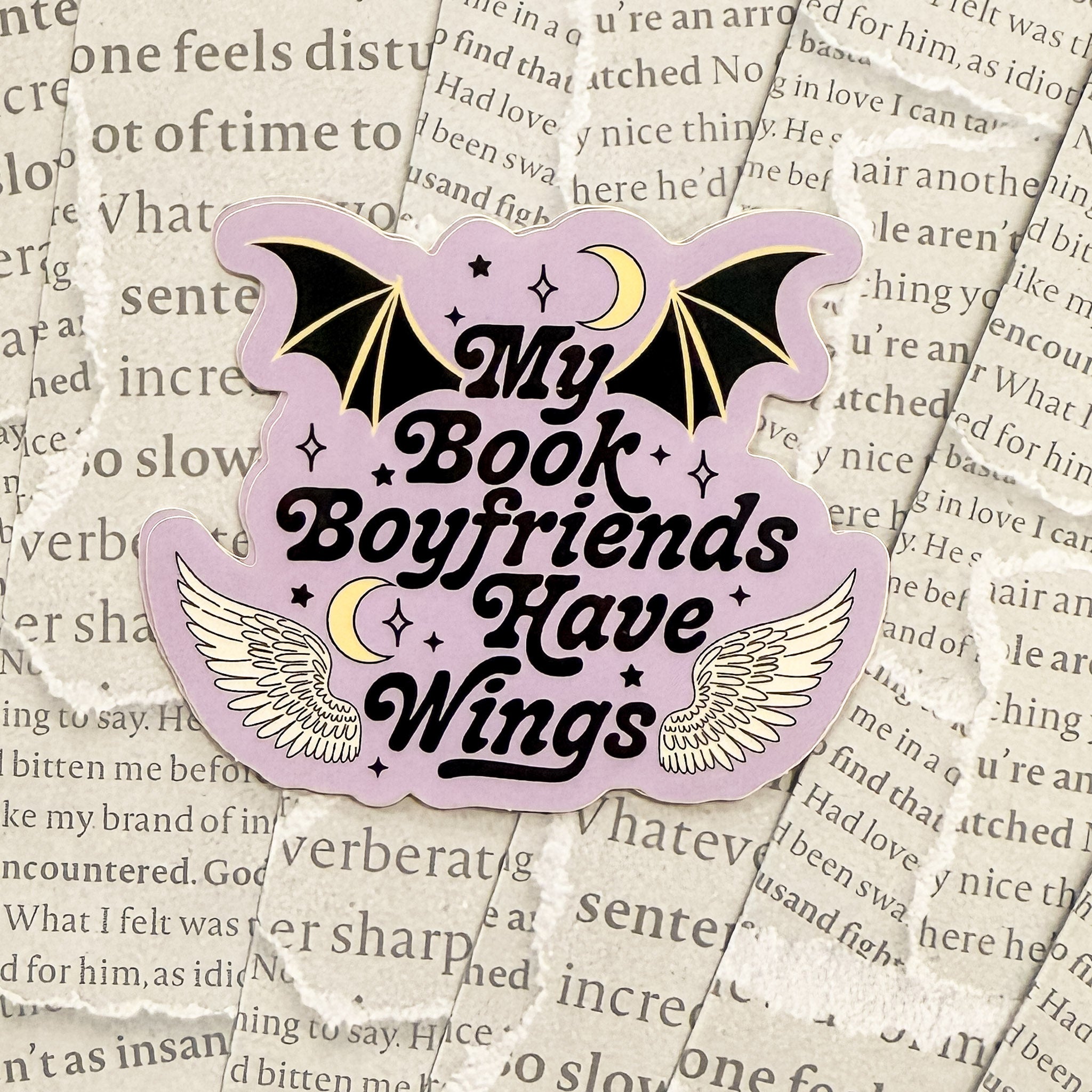 purple vinyl sticker that says "my book boyfriends have wings" with bat wings, stars and a crescent moon, on a background made of torn black and white book pages