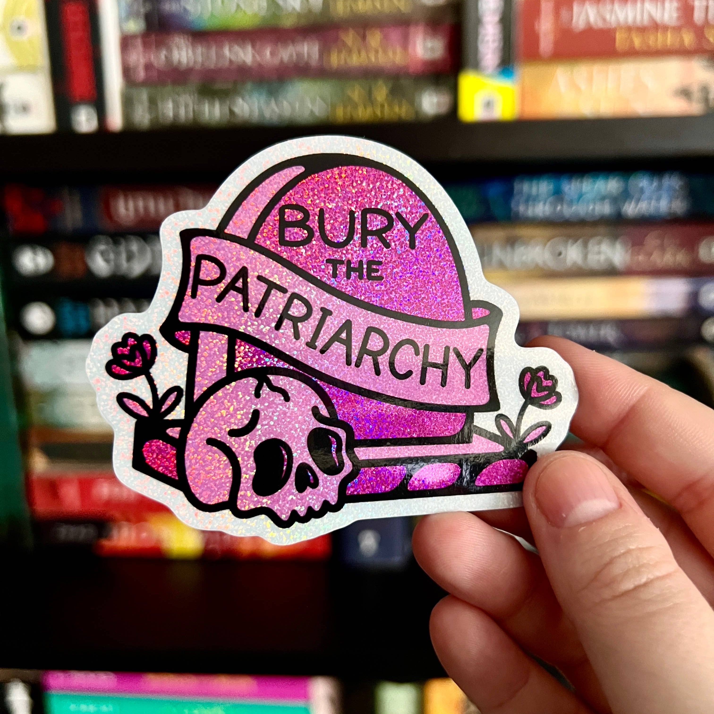 Holographic sticker of a pink gravestone that reads “Bury the Patriarchy” with a skull and flowers.
