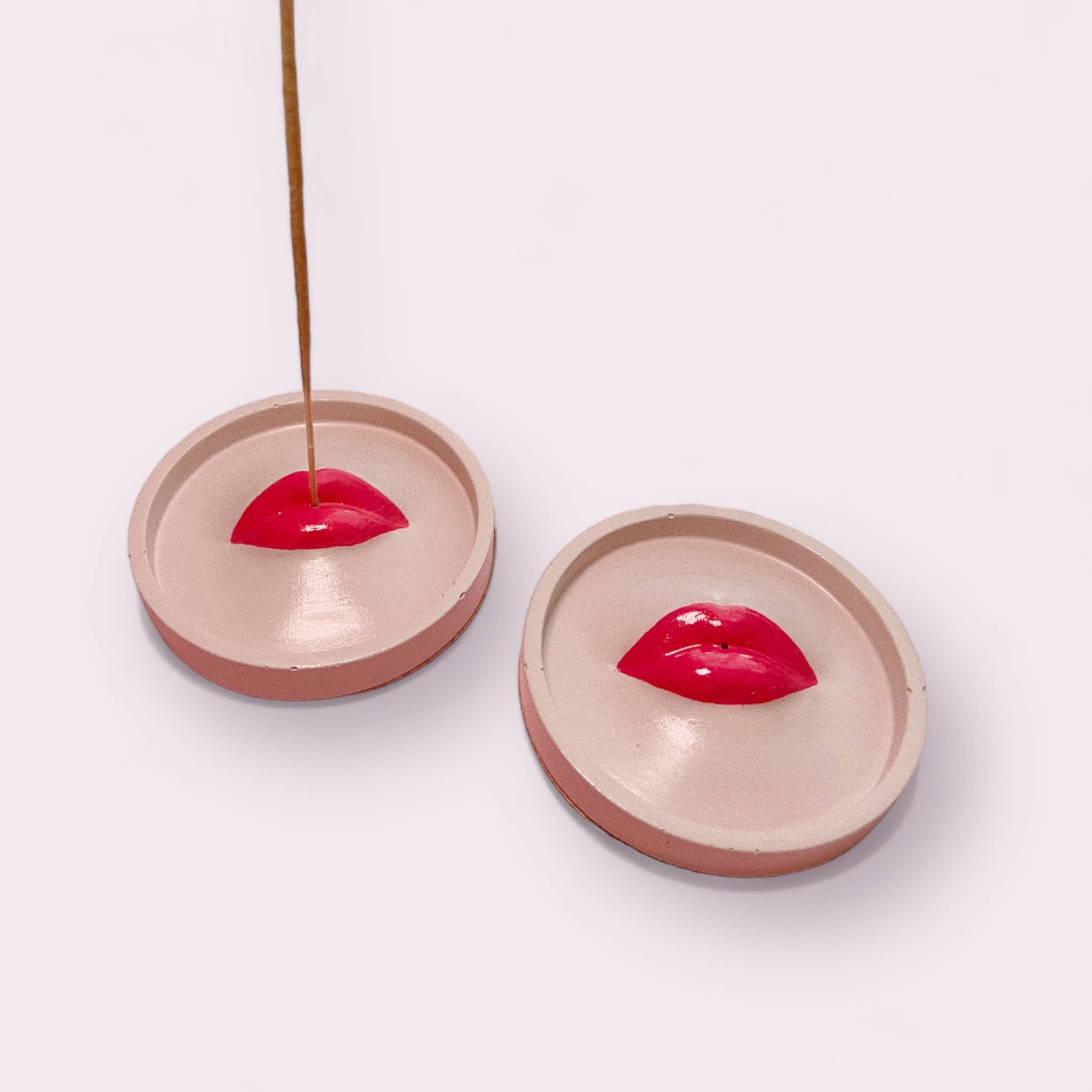 Handmade concrete incense holders with glossy red lips in the center, used to hold incense sticks. Set on a light pink background