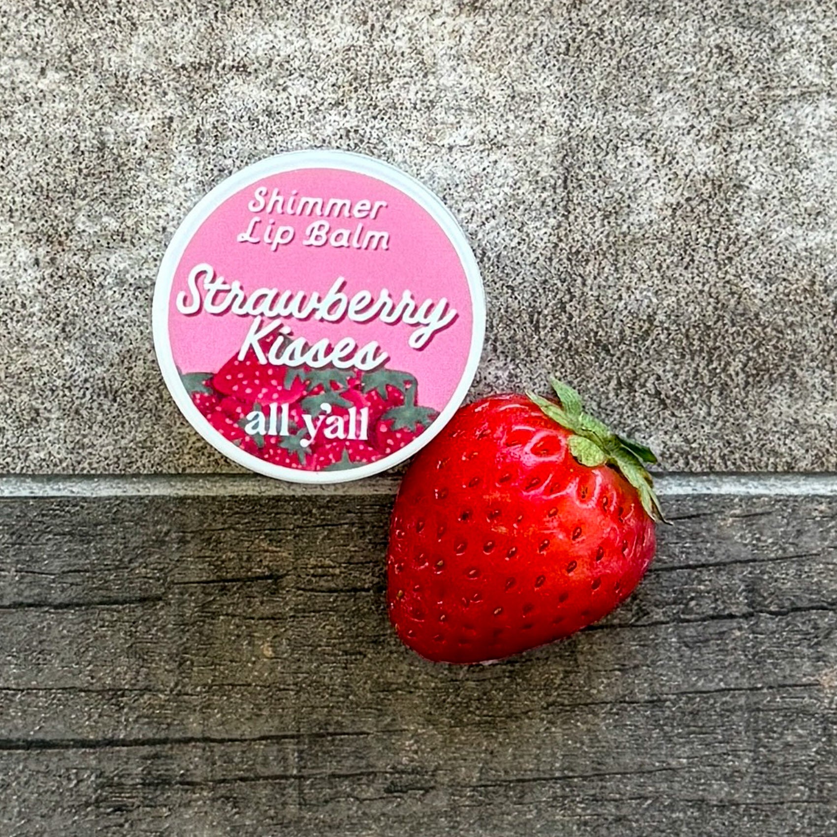 Round lip balm tin labeled “Strawberry Kisses” next to a fresh strawberry. Tinted shimmer balm inspired by Cowboy Casual.