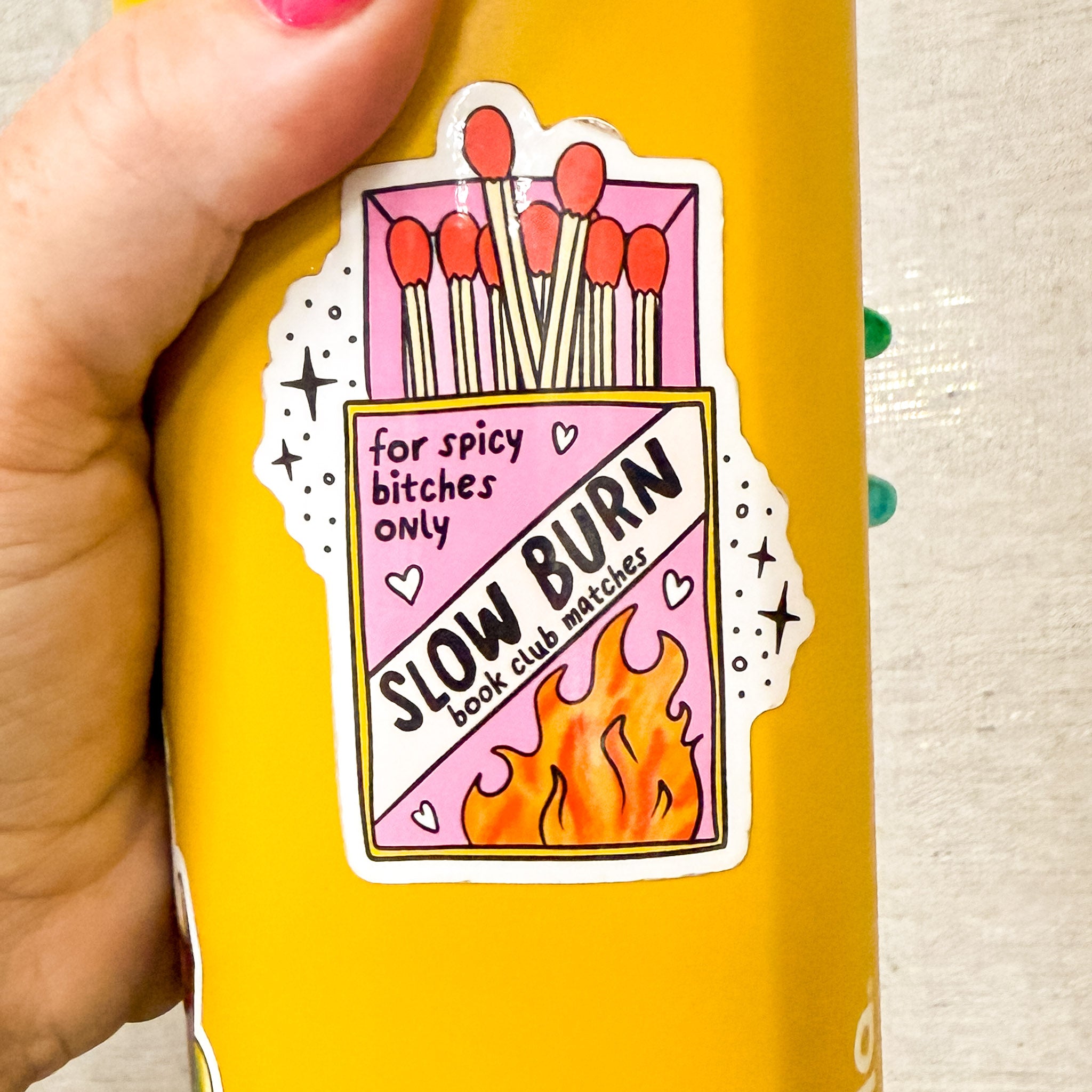 A vinyl sticker shaped like a pink and yellow matchbox labeled “Slow Burn book club matches – for spicy [censored] only,” with flames and hearts, on mustard green owalla bottle