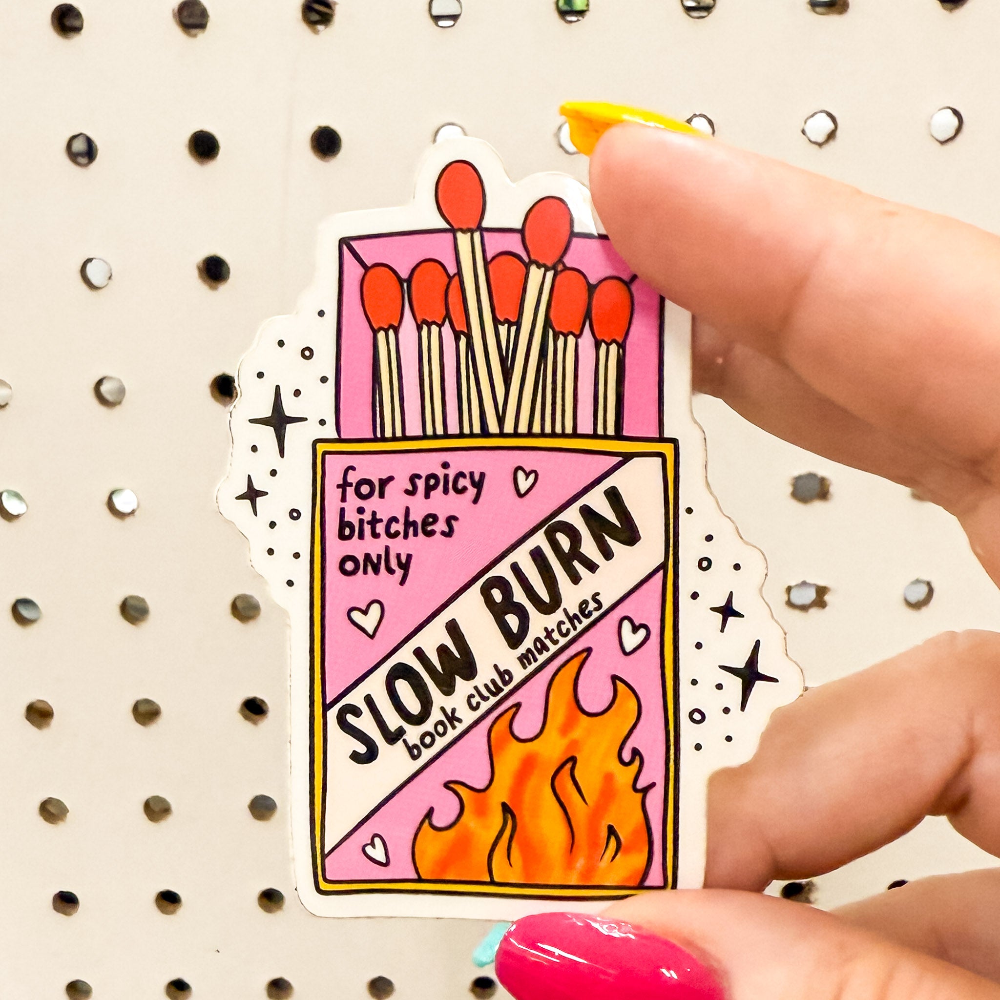 A vinyl sticker shaped like a pink and yellow matchbox labeled “Slow Burn book club matches – for spicy [censored] only,” held in hands with rainbow nails, on textured background.
